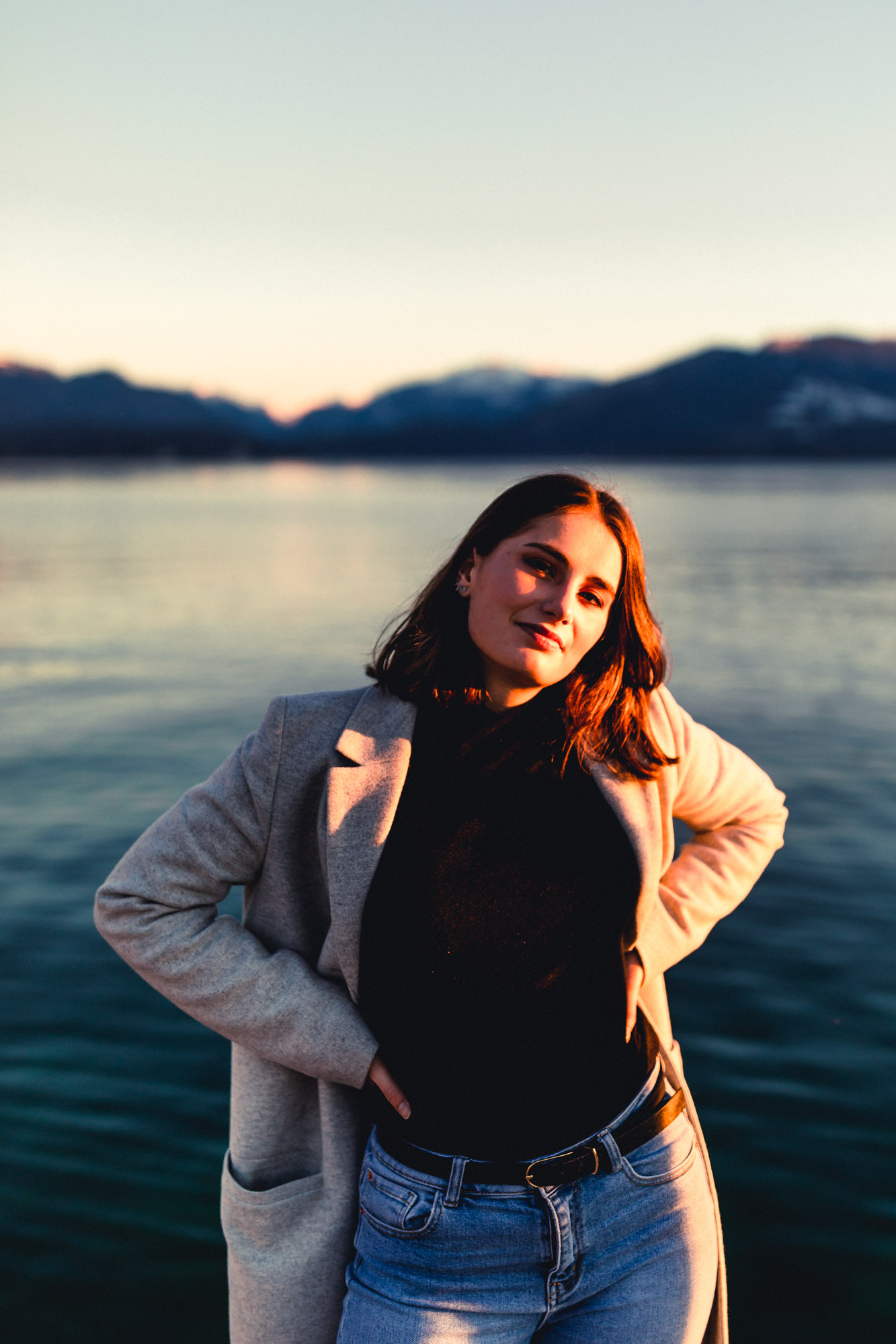 photo d'une femme, mains sur les hanches avec une veste grise, un haut noir et un jeans bleu, lumière du coucher de soleil. elle se tient debout devant de l'eau et des montagnes