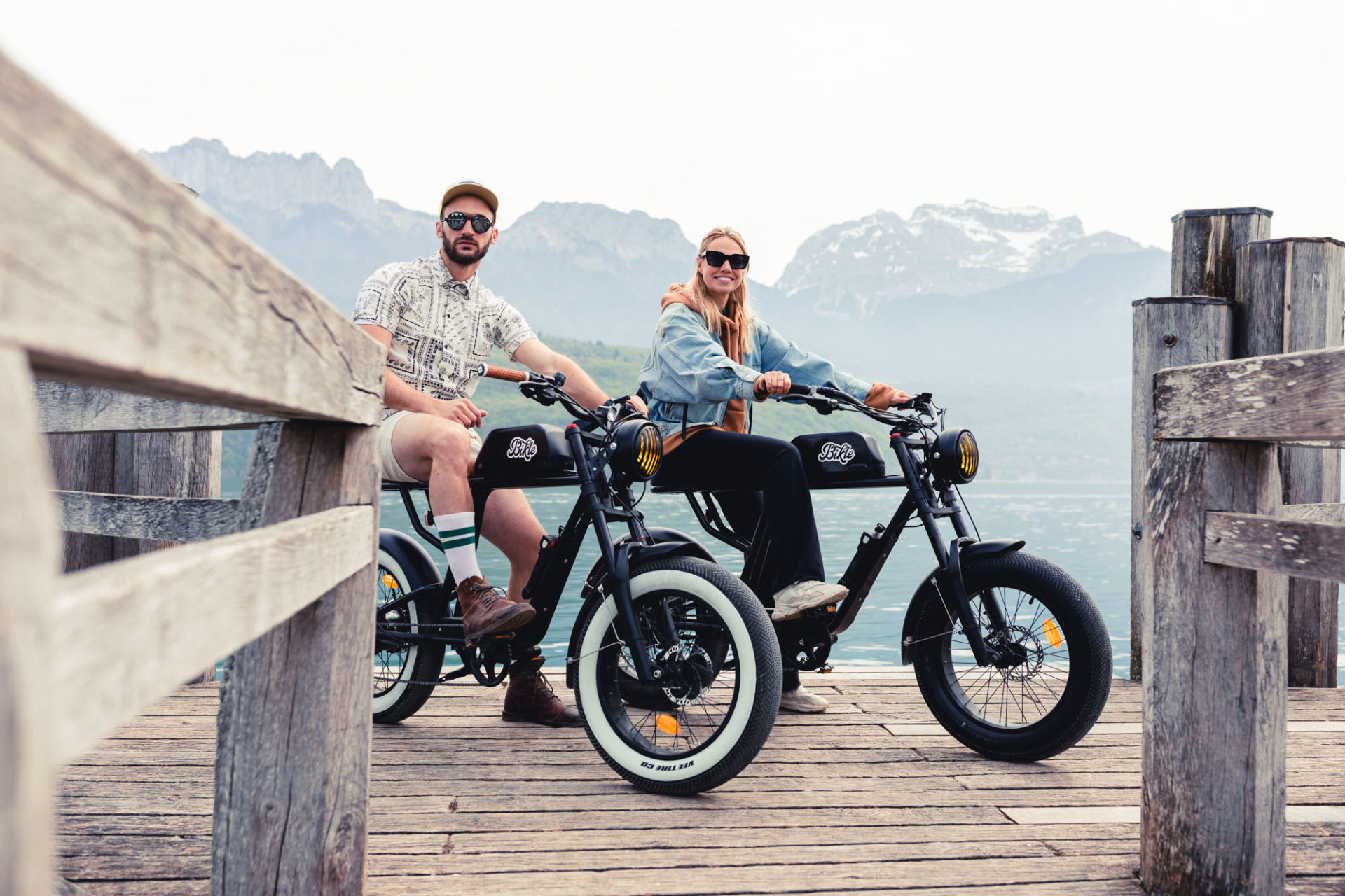 photo de deux personnes assises sur deux vélos électrique noirs ressemblant à une vieille moto, devant des montagne et un lac, sur un ponton en bois