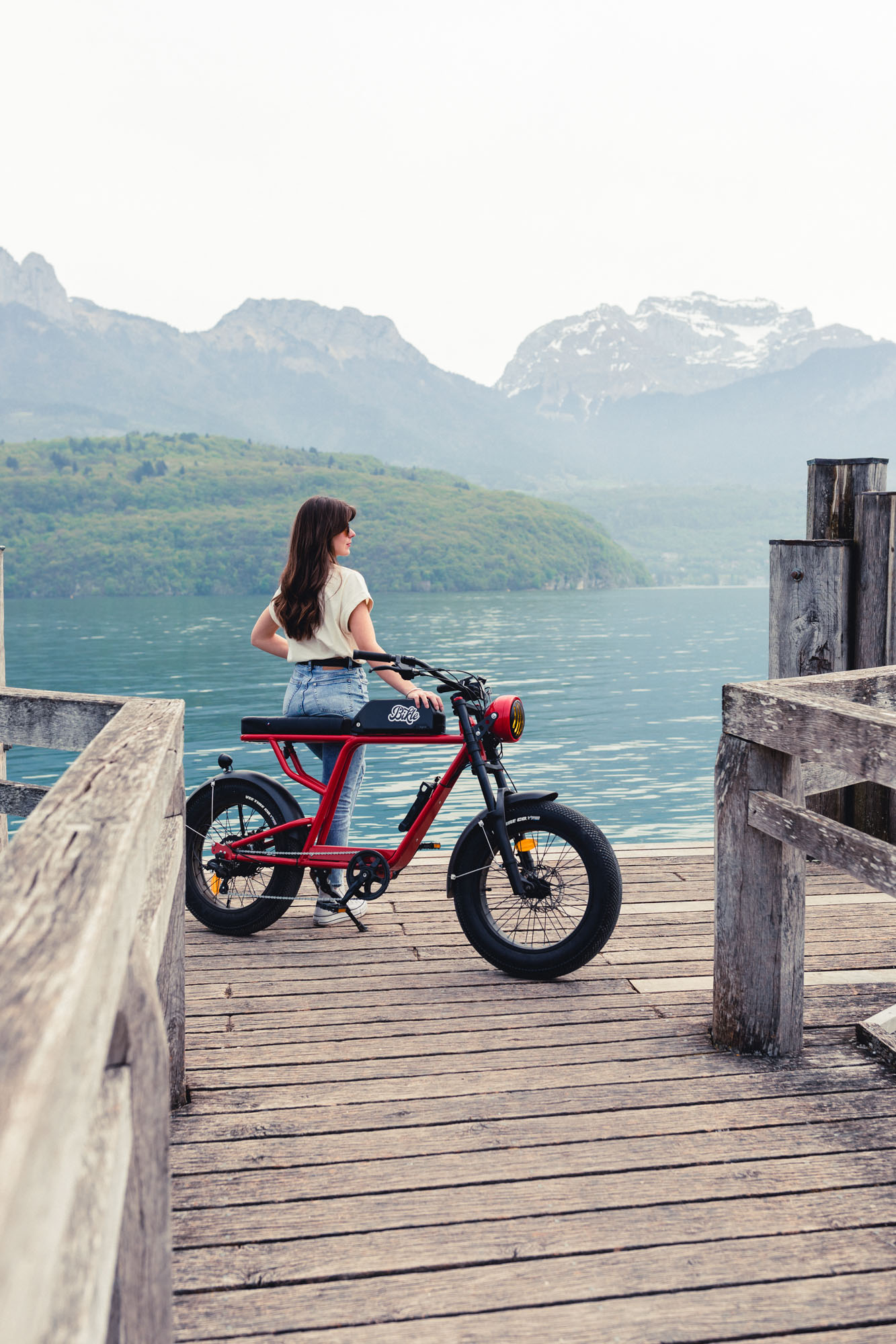 photo d'une femme brune cheveux longs tenant un vélo électrique rouge ressemblant à une vieille moto, devant des montagne et un lac, sur un ponton en bois