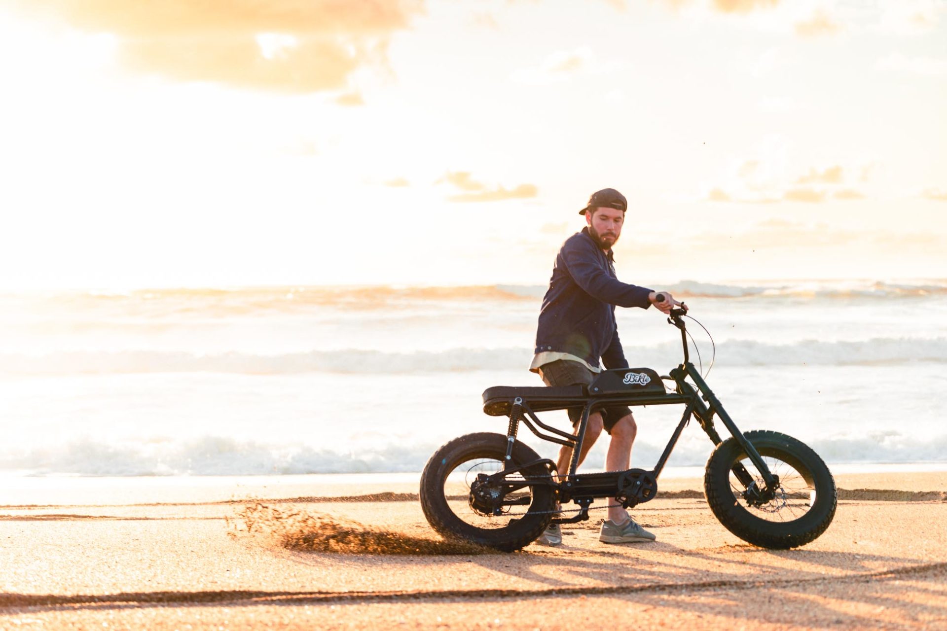 Photo d'un homme tenant un vélo électrique faisant des dérapages sur une plage au bord de la mer.