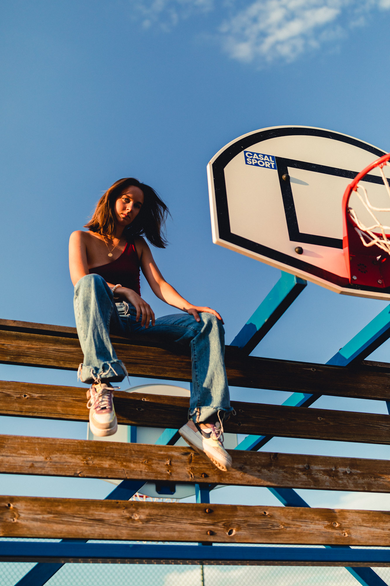 Photo d'une femme blonde assise à côté d'un panier de basketball, habillée en débardeur rouge, jeans bleu et chaussures Nike.