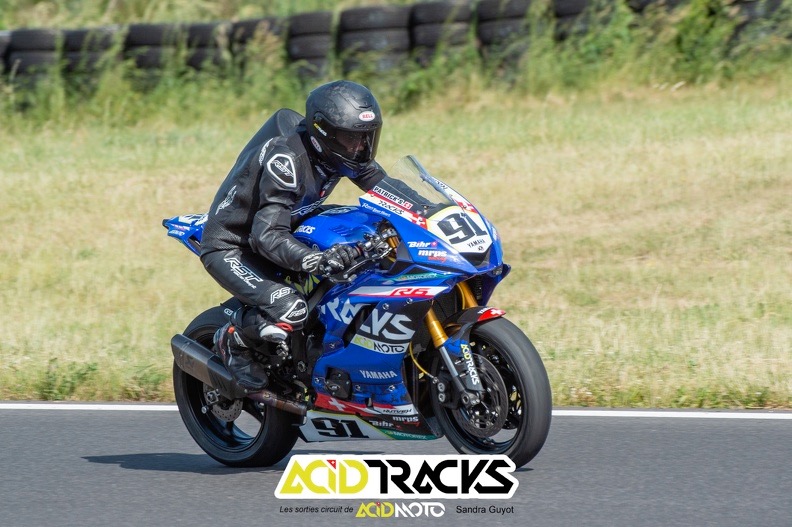 Photo d'une moto bleue en mouvement, floquée AcidMoto et Acidtracks, avec pilote en combinaison noire.
